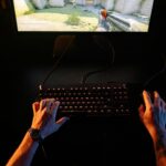 A gamer intensely playing a first-person shooter game on a computer setup indoors at night.